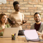 Young, diverse group of professionals working at a conference table with a brick background, illustrating the blog topic of Big Benefits for Small Teams: Health Insurance Options that Work
