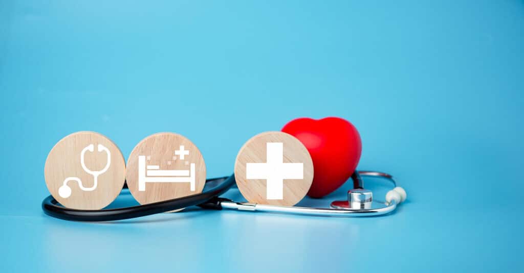 Stethoscope and red heart on blue background with circular wooden blocks showing health care icons, illustrating the blog topic "Private vs Public Health Insurance: What You Think You’re Buying… and What You Actually Are"